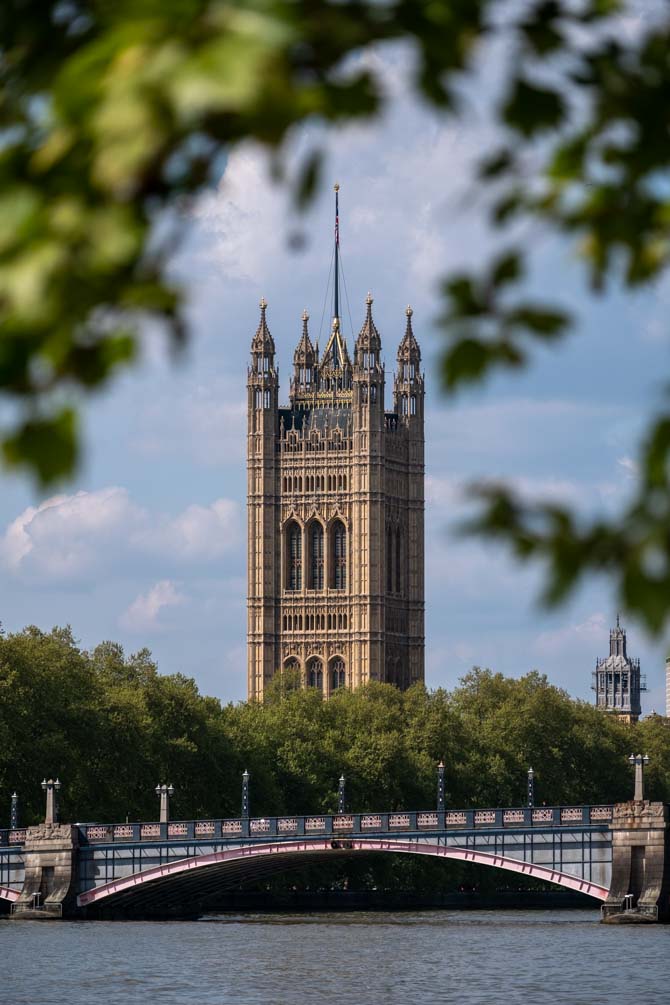 Victoria Tower, of the houses of Parliament, with a bridge in front and trees framing the shot, viewed from the south bank of the Thames Path.